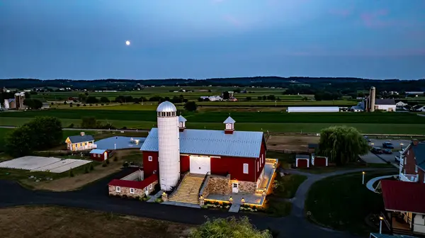 stock image Ronks, Pennsylvania, USA, July 19 2024 - Serene Farm Scene Dusk Featuring Large Red Barn With Silo, Stone Courtyard Illuminated By Soft Lighting, Charming Stone House Reflecting In Pond, Expansive