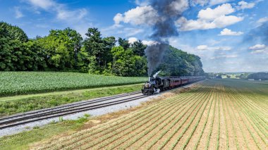 Ronks, Pensilvanya, ABD, 8 Haziran 2024 - Siyah Buharlı Lokomotif Kırmızı Yolcu Arabalarını Dolgun Yeşil Araziler ve Ormanlık Alanlarda, Açık Mavi Bulutlar Altında Çekiyor