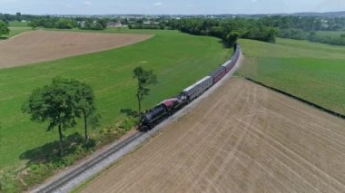 Ronks, Pennsylvania, ABD, 27 Mayıs 2022 - Yeşil ve kahverengi tarlalarda yol alan bir trenin havadan görünüşü, çayırları noktalayan ağaçlar, paralel giden bir toprak yol ve A 'nın altındaki uzak bir kasaba