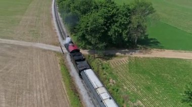 Ronks, Pensilvanya, ABD, 10 Haziran 2022 - Kırmızı çatılı bir trenin her iki tarafında yeşil ve kahverengi alanlar bulunan ve A altında duman ve buhar yayan havadan görünüşü