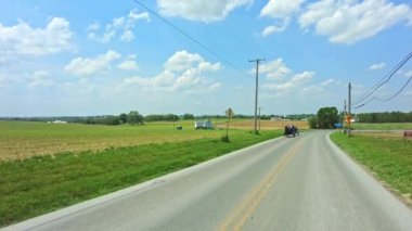 Ronks, Pennsylvania, ABD, 22 Mayıs 2022 - Çevresi yeşil alanlar, elektrik hatları ve tüylü açık mavi gökyüzü ile çevrili virajlı kırsal yolda seyahat eden yolcuları taşıyan iki at arabası