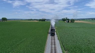 Ronks, Pennsylvania, ABD, 28 Haziran 2022 - Uçsuz bucaksız, parlak mavi gökyüzü altındaki yeşil tarım arazileri boyunca kırmızı çatılı bir trenle küçük bir çiftlik evinin önünden geçen