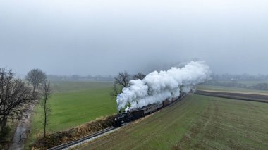 Bir buhar treni yeşil tarlalardan geçip sisli havaya duman bulutları salıyor ve raylar boyunca ilerliyor..