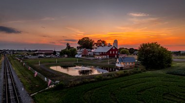 Ronks, Pennsylvania, ABD, 19 Temmuz 2024 - Günbatımındaki Huzurlu Çiftlik Sahnesi Red Barn, Silos, Pond, Çevresindeki Yeşil Alanlar, Ön plandaki Tren Rayları Dramatik, Renkli Gökyüzü Aydınlatıcı