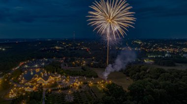 Aydınlatılmış binalarla dolu, parlak bir fuar alanı üzerinde beyaz havai fişekler patlıyor ve Ufukta şehrin ışıkları parıldıyor..