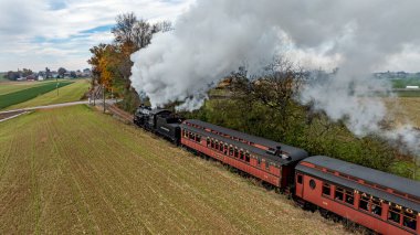 Ronks, Pennsylvania, ABD, 4 Ekim 2023 - Bir buhar treni, sonbaharda ağaçlar ve tarlalar arasında dalgalanan dumanla kırsal bir hat boyunca tıkırdıyor.