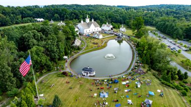 Elizabethtown, Pennsylvania, ABD, 4 Temmuz 2024 - Serene Pond, Beyaz Ahırlar, Küçük Kiliseler ve Yeşil Ağaçlar ve Yuvarlanan Tepeler ile çevrelenmiş insanların eğlendiği büyüleyici kırsal panayır