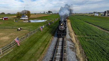 Bir buhar treni yeşil bir kırsalda demiryolu boyunca tıkırdıyor, açık gökyüzünün altındaki bayrakları ve tarlaları geçiyor..