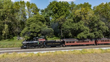 Ronks, Pennsylvania, ABD, 4 Ekim 2023 - Buharlı bir lokomotif, bulutsuz bir gökyüzünün altında etrafını çevreleyen canlı ağaçlarla çevrili bir yolcu vagonunu çekiyor.