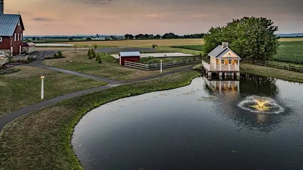 stock image Ronks, Pennsylvania, USA, July 19 2024 - Charming Farm Scene At Dusk Featuring A Small Stone House With A White Porch Reflecting In A Pond, Surrounded By Green Fields, Fenced Pastures, And A Softly