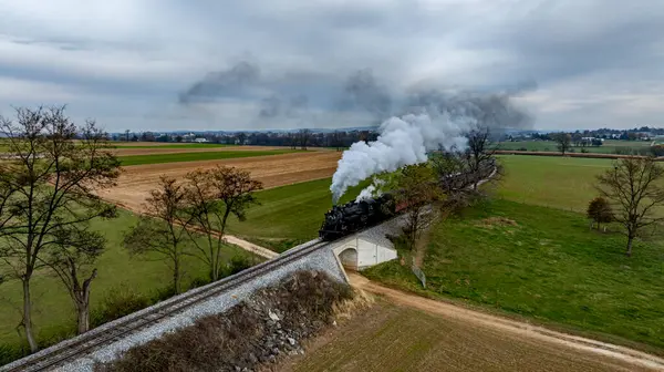 stock image A vintage steam train chugs along tracks, releasing smoke, while passing through green fields amid overcast skies.