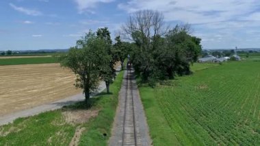 Kırmızı çatısı olan buharlı bir tren, yemyeşil tarlalardan geçen ve hasat edilen arazilerden geçen, güneşli bir günde uzun ağaçların altından geçen, uzak bir yerdeki çiftlik binaları olan bir pist boyunca ilerliyor.