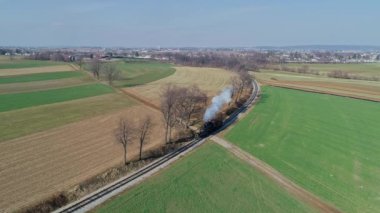 Ronks, Pennsylvania, ABD, 11 Mart 2022 - Parlak mavi bir gökyüzü altında verimli tarlalar ve ağaçlar arasında bir buhar treni tıkırdıyor. Yüksek kalite 4k görüntü