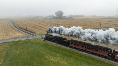 Eski bir buhar treni, gri bir gökyüzü altında geniş tarlalar ve uzak çiftlik yapılarıyla çevrili, sisli kırsal bir ortamdan geçerken kalın duman bulutları püskürtür..