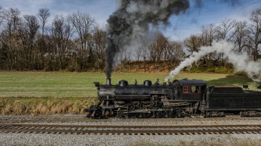 Buharlı bir lokomotif, manzaralı bir kırsal bölgede tren rayları boyunca ilerlerken duman bulutları yayar. Canlı doğal çevre, sonbahar yapraklarının güzelliğini vurguluyor..