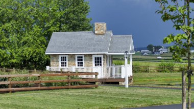 Açık mavi gökyüzü ve birkaç bulutla yemyeşil bir tarlada duran tuhaf bir taş kulübe. Geniş arazide ahşap bir çit ve manzaralı bir manzara var..