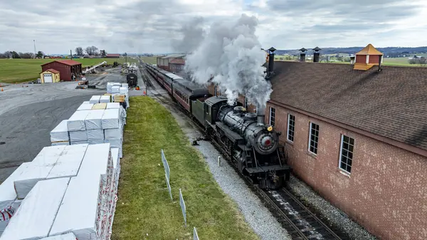 Stock image A vintage steam locomotive emits steam while departing from a brick station. Nearby, warehouses and freshly stacked materials are visible, creating a historic railroad atmosphere.