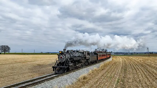 Eski bir buhar treni tren rayları boyunca tıka basa doluyor. Gri, bulutlu bir gökyüzü altında toplanan altın mahsul tarlalarından geçerken buhar bulutları salıyor..