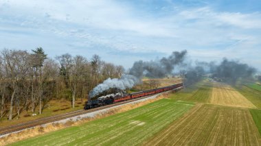 Buharlı bir lokomotif, parlak bir gökyüzünün altındaki ağaçların ve tarlaların üzerine duman ve buhar saçan antika bir treni güzel bir kırsala çeker..