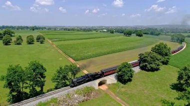 Ronks, Pennsylvania, ABD, 19 Temmuz 2022 - Pahalı bir buhar treni canlı yeşil tarlalar ve mavi gökyüzü üzerinde seyahat ederek kırsal yaşamın güzelliğini ve yüksek kaliteli 4k 'yi gözler önüne seriyor.