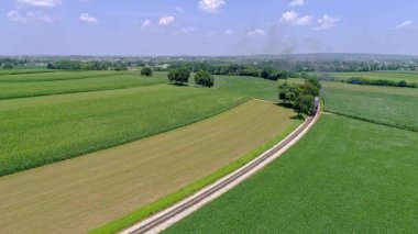 Bir tren, açık mavi bir gökyüzünün altında canlı yeşil tarlalarla çevrili dolambaçlı bir yol boyunca hareket eder. Kırsal manzara, huzurlu bir kırsal alanda yazın özünü yakalar. - Evet. Yüksek kalite 4k