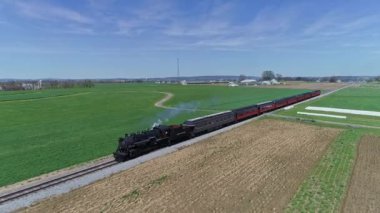 Ronks, Pennsylvania, ABD 20 Nisan 2022 - Lancaster County 'nin yemyeşil tarlalarında bir buhar treni süzülerek temiz mavi bir gökyüzü altında kırsal alanı gösteriyor. - Evet. Yüksek kalite 4k görüntü