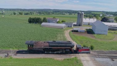 Ronks, Pennsylvania, ABD 28 Haziran 2022 - Parlak mavi gökyüzü altında yemyeşil tarlalar ve çiftlik binalarıyla çevrili raylar boyunca tamir edilen eski bir buhar lokomotifi. - Evet. Yüksek