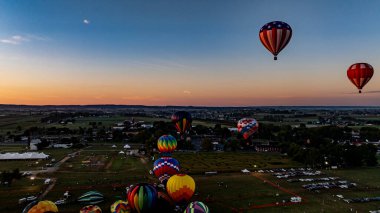 Sıcak hava balonları alacakaranlık gökyüzüne fırlatılıyor, kırsal alanların ve şenliklerin olduğu görkemli bir kırsal alanda sönen ışığa karşı canlı bir ekran oluşturuyor..