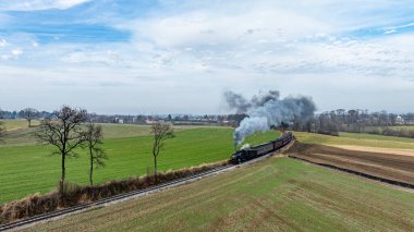 Geniş yeşil alanlar ve uzak ağaçlarla çevrili tren rayları boyunca uzanan buhar lokomotifi bulutlu gökyüzüne doğru bir duman bulutu yayarak sakin bir kırsal alan oluşturur..