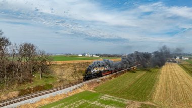 Klasik bir buhar lokomotifi, mavi gökyüzü altında geniş tarım arazileri boyunca hareket eden duman bulutları yayarak demiryolu rayları boyunca seyahat eder. Uzak ahırlar ve silolar manzarayı noktalıyor.