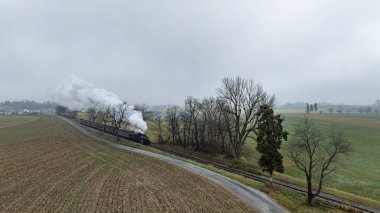 Antika bir buhar treni bulutlu ve sisli bir gökyüzünün altında tarlalar ve ağaçlarla çevrili demiryolu rayları boyunca süzülür. Manzara, sabahın erken saatlerinde sakin kırsal bir çevreyi gözler önüne seriyor..