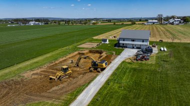 Ağır makineler, bir çiftlik evinin yakınındaki inşaat sahasında çalışıyor. Arazi parlak gökyüzü ve geniş tarlalar altında yeni gelişmeler için hazırlanıyor..