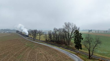 Bir buhar treni, çıplak ağaçlar ve bulutlu bir gökyüzünün altındaki yuvarlanan tarlalarla birlikte sessiz bir kırsalda ilerlerken beyaz bir duman çıkarır ve sabahın erken saatlerinde nostaljik bir atmosfer yaratır..