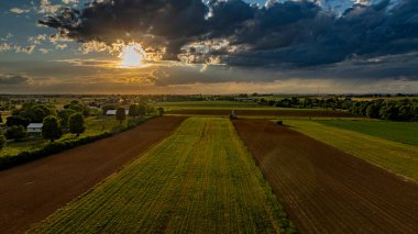 Güneş canlı bir gökyüzünde batarken, geniş tarım alanları, ağaçlar ve çiftlik yapılarıyla kaplı, sakin ve resimsel kırsal bir manzara yaratıyor..