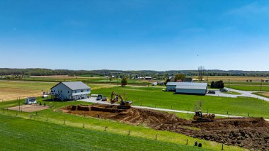 Ağır makineler, açık mavi gökyüzünün altındaki tarlalarla çevrili bir evin yakınındaki inşaat sahasında çalışıyor. Bölge hareketlilikle dolup taşıyor, bu da yeni gelişim sürecinde ilerleme olduğunu gösteriyor..