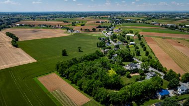 Geniş bir hava manzarası yeşil tarlaların, kahverengi tarım arazilerinin ve güneşli bir günde yeşilliklerin arasına yuva yapmış küçük bir topluluğun yer aldığı canlı bir kırsal alan yakalar..