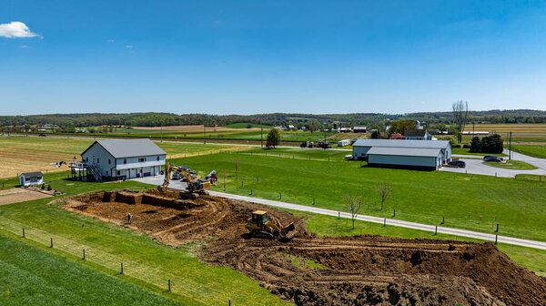 Тяжелая техника работает на строительной площадке рядом с домом, окруженной полями под ясным голубым небом. Область кипит активностью, указывая на прогресс в новом развитии.