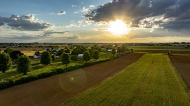 Güneş canlı bir gökyüzünde batarken, geniş tarım alanları, ağaçlar ve çiftlik yapılarıyla kaplı, sakin ve resimsel kırsal bir manzara yaratıyor..