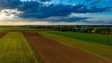 Güneş canlı bir gökyüzünde batarken, geniş tarım alanları, ağaçlar ve çiftlik yapılarıyla kaplı, sakin ve resimsel kırsal bir manzara yaratıyor..