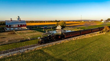Ronks, Pennsylvania, ABD, 20 Ekim 2024 - Tarihsel buharlı tren raylar boyunca hareket ederek, geniş tarım arazileri oluşturdu. Güneş batıyor, sıcacık altın renklerini yeryüzüne saçıyor.,