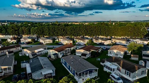 Hava görüntüsü, uzun ağaçlarla çevrili Mobil, Prefabrik, Üretilmiş Evler Mahallesi 'ni yakalıyor. Güneş arka planda batıyor, çatıların üzerinde ılık bir parıltı bırakıyor..