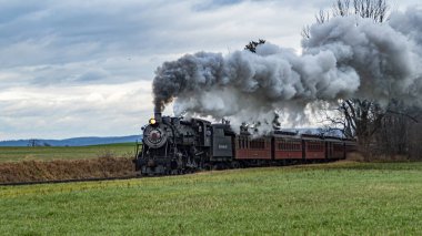 Kaliteli bir buhar treni engin kırsal alandaki tren raylarını tıka basa doldurur ve kara bulutlara yoğun duman bulutları yayar. Yakın tarlalarda altın mahsuller sergileniyor.