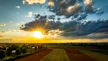 A vibrant sunset casts warm colors over expansive farmland, with lush green fields and patches of clouds creating a picturesque sky. The serene landscape reflects the tranquility of rural life. clipart