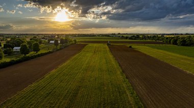 Güneş canlı bir gökyüzünde batarken, geniş tarım alanları, ağaçlar ve çiftlik yapılarıyla kaplı, sakin ve resimsel kırsal bir manzara yaratıyor..