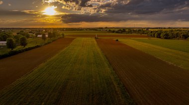 Canlı bir kır manzarası dramatik bir gökyüzü altında geniş yeşil ve kahverengi tonlarını sergiliyor. Güneş batıyor, tarım arazisine ve huzurlu çevreye sıcak bir parıltı saçıyor..