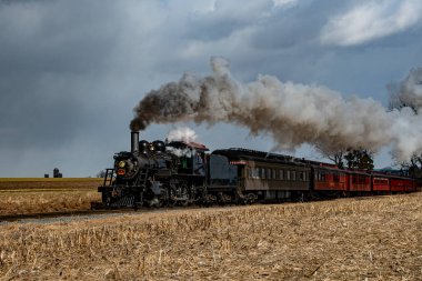 Kaliteli bir buhar treni engin kırsal alandaki tren raylarını tıka basa doldurur ve kara bulutlara yoğun duman bulutları yayar. Yakın tarlalarda altın mahsuller sergileniyor.