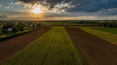 Güneş canlı bir gökyüzünde batarken, geniş tarım alanları, ağaçlar ve çiftlik yapılarıyla kaplı, sakin ve resimsel kırsal bir manzara yaratıyor..