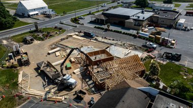 Ticari bir konumda çalışan işçilerin yeni bir binayı aktif olarak çerçeveletmesiyle bir inşaat projesi ilerliyor. Yapının ahşap çerçeveleri var ve açık bir gökyüzü ve yeşil manzaranın ortasında yer alıyor..