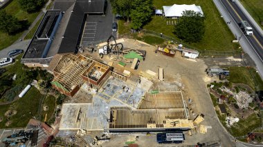 Ticari bir konumda çalışan işçilerin yeni bir binayı aktif olarak çerçeveletmesiyle bir inşaat projesi ilerliyor. Yapının ahşap çerçeveleri var ve açık bir gökyüzü ve yeşil manzaranın ortasında yer alıyor..