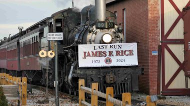 Strasburg, Pennsylvania, December 31, 2024 - A vintage steam train travels through a rural landscape, displaying a sign in tribute to James R. Rice during its final journey, capturing a moment of nostalgia and celebration. clipart